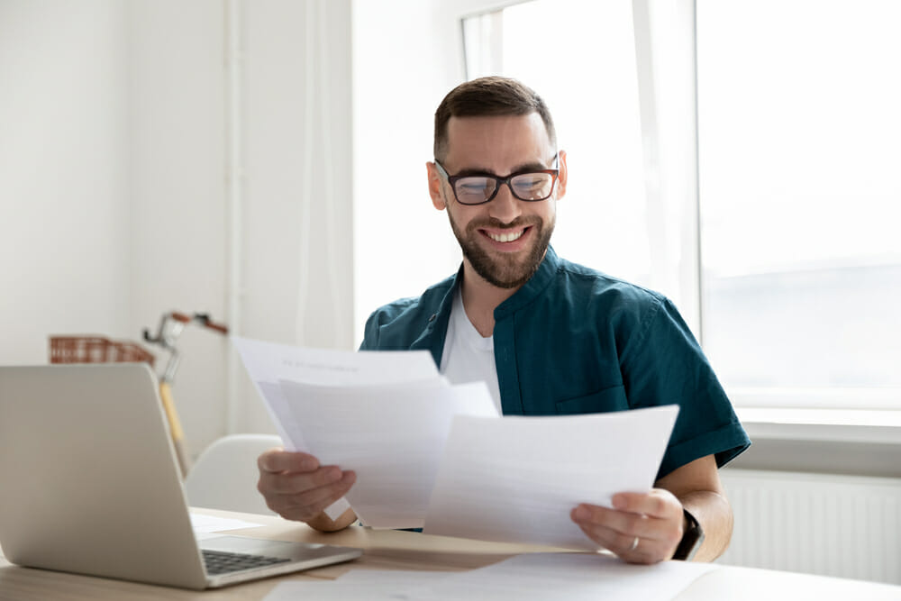 Man sitting at a table in front of his laptop holding papers in front of him